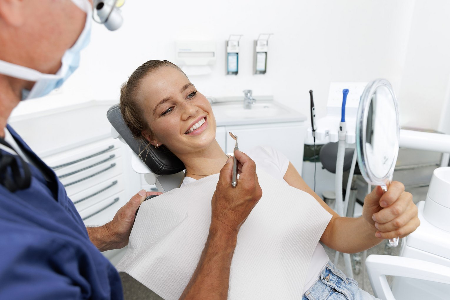 Patientin liegt auf dem Behandlungsstuhl und lächelt. Sie hält einen Spiegel in der Hand. Der Arzt beginnt mit der Behandlung.